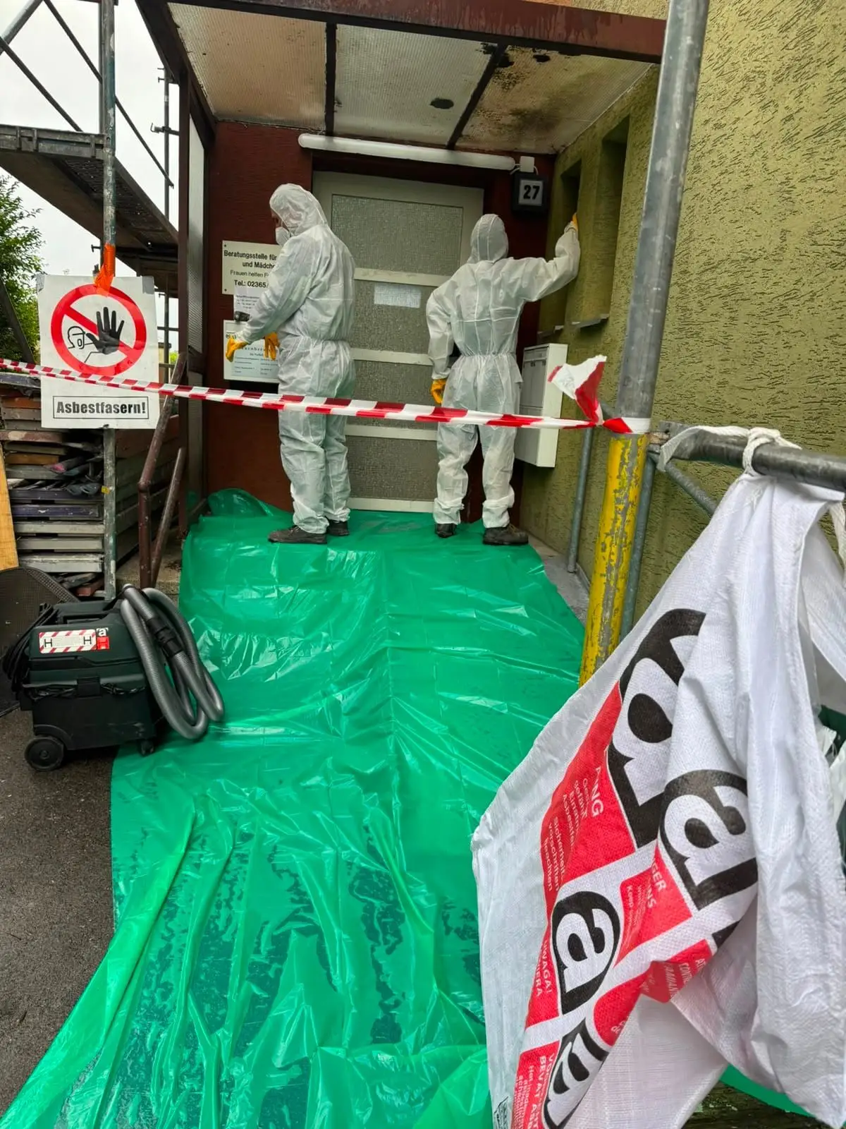 Fachkräfte im Schutzanzug am abgeschotteten Zugang zum Arbeitsbereich – Abschottungsfolie, Warnschild „Asbestfasern" und Big-Bags für die Entsorgung