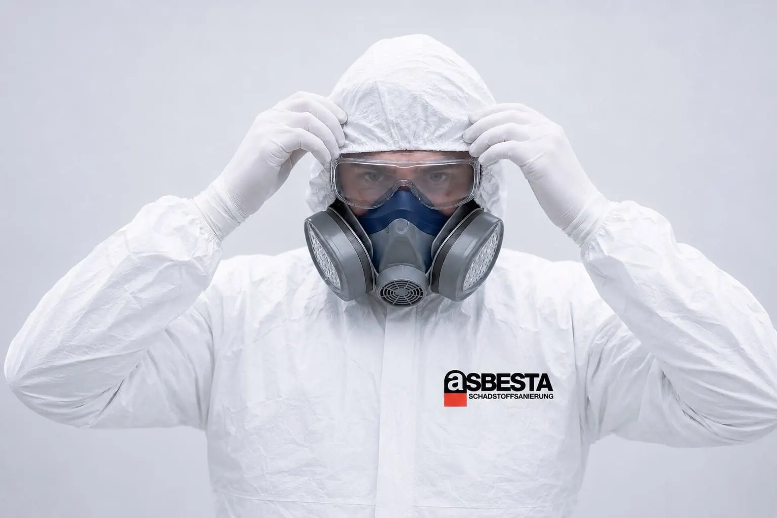 Fachkraft von Asbesta im weißen Einweg-Schutzanzug legt Halbmaske und Schutzbrille an – ASBESTA-Logo auf der Brust